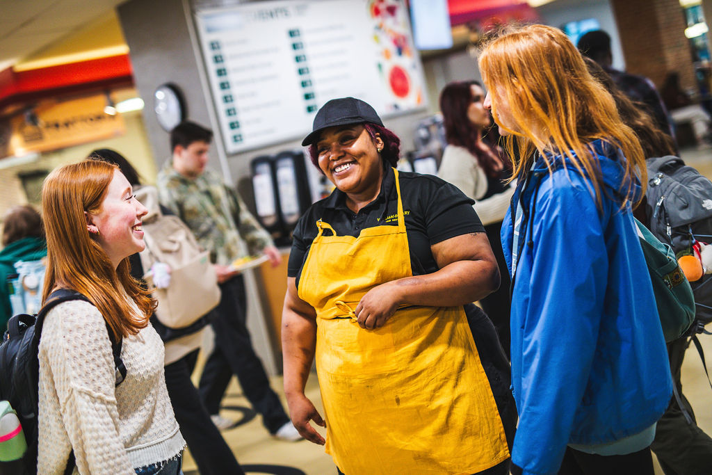 A W&M Dining employee in a golden apron smiles and laughs with two students