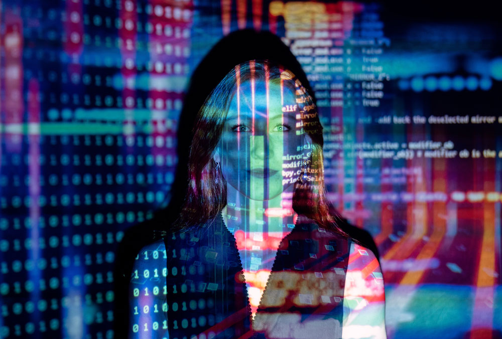 Woman standing against a dark background with colorful lines of computer code, binary numbers, and data visualizations projected across her face and body.
