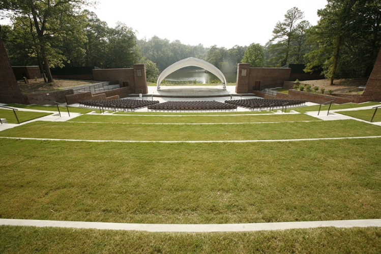 Large tiered amphitheater with grass for further away from stage and seats closer to stage. stage is covered by large tent with a lake in the background.