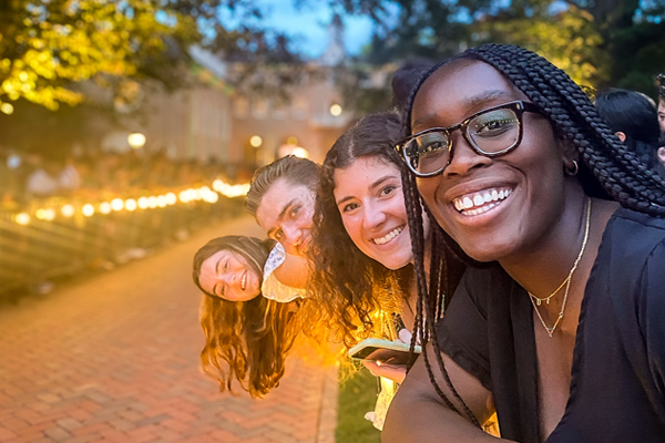 Four smiling students leaning into view around a lit brick walkway